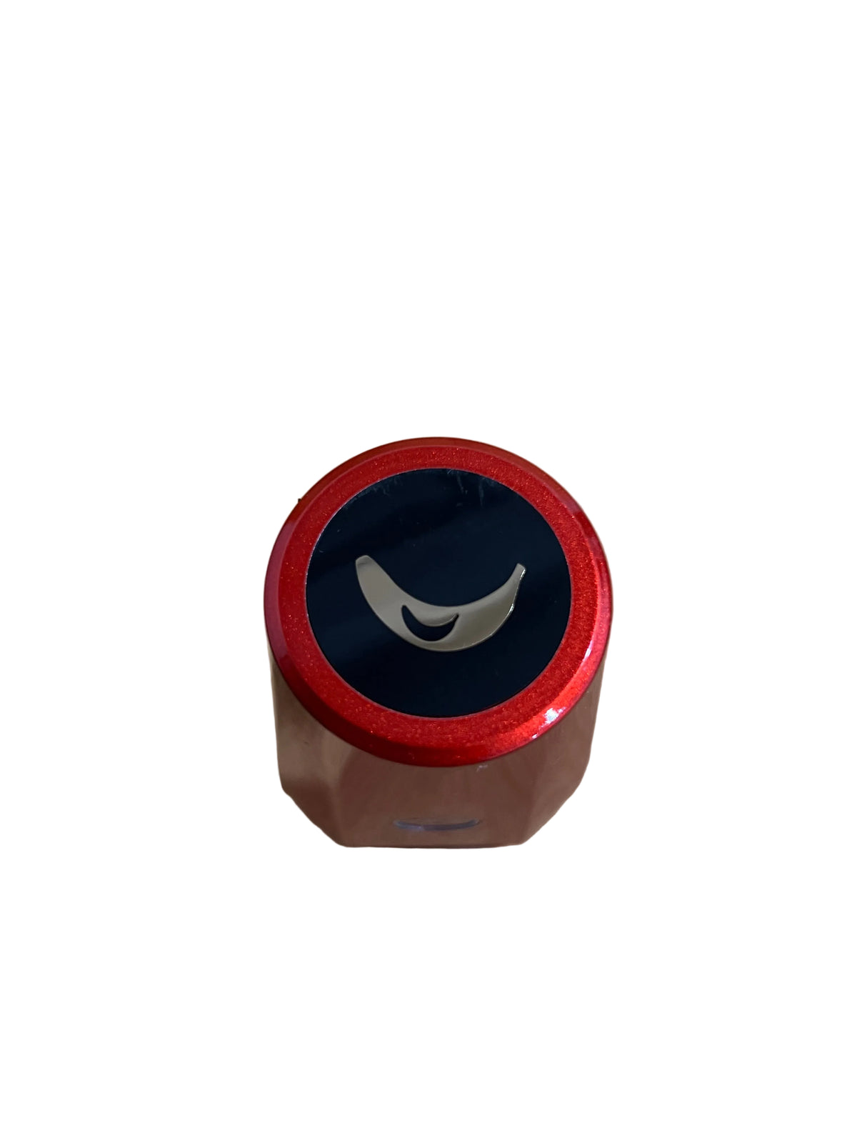 A close-up top-down view of a small cylindrical product with a metallic red rim, a dark navy blue center, and a white curved logo, placed on a beige textured surface.